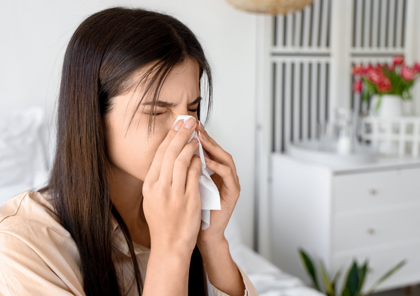 Woman sitting on her bed blowing her nose into a tissue in a bright bedroom
