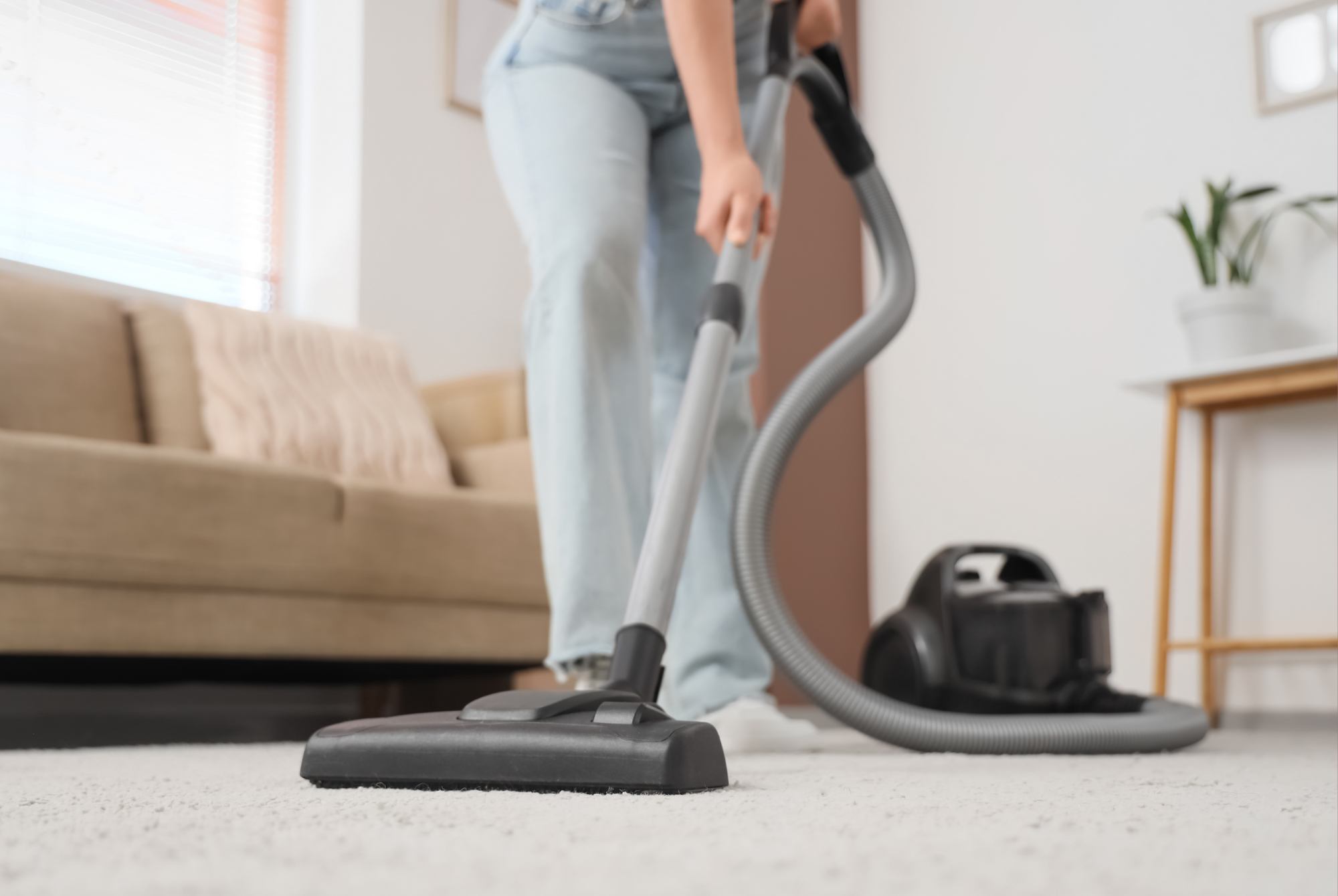 A person vacuuming a light-colored carpet in a living room beside a beige sofa